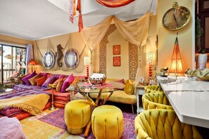 Ein lebendiges Wohnzimmer mit einer Couch, Poufs, einem Glastisch und einem Essbereich mit weißem Tisch.