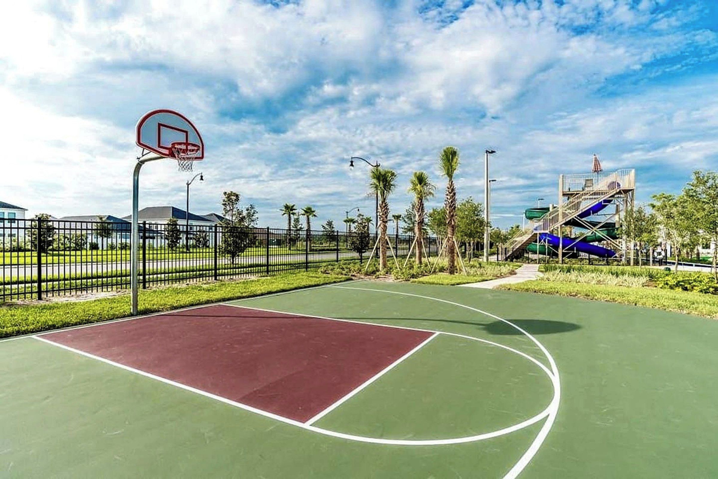 An outdoor basketball court with a red playing surface, surrounded by a fence, palm trees, and a water slide structure.