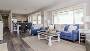 A modern living room with a blue sofa, a coffee table, and a dining area with a table and chairs. - Stunning 3 Bedroom Condo Near Crystal Pier (Wrightsville Beach)