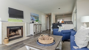 A living room with a fireplace, blue sofas, a coffee table, and a kitchen in the background. - Stunning 3 Bedroom Condo Near Crystal Pier (Wrightsville Beach)