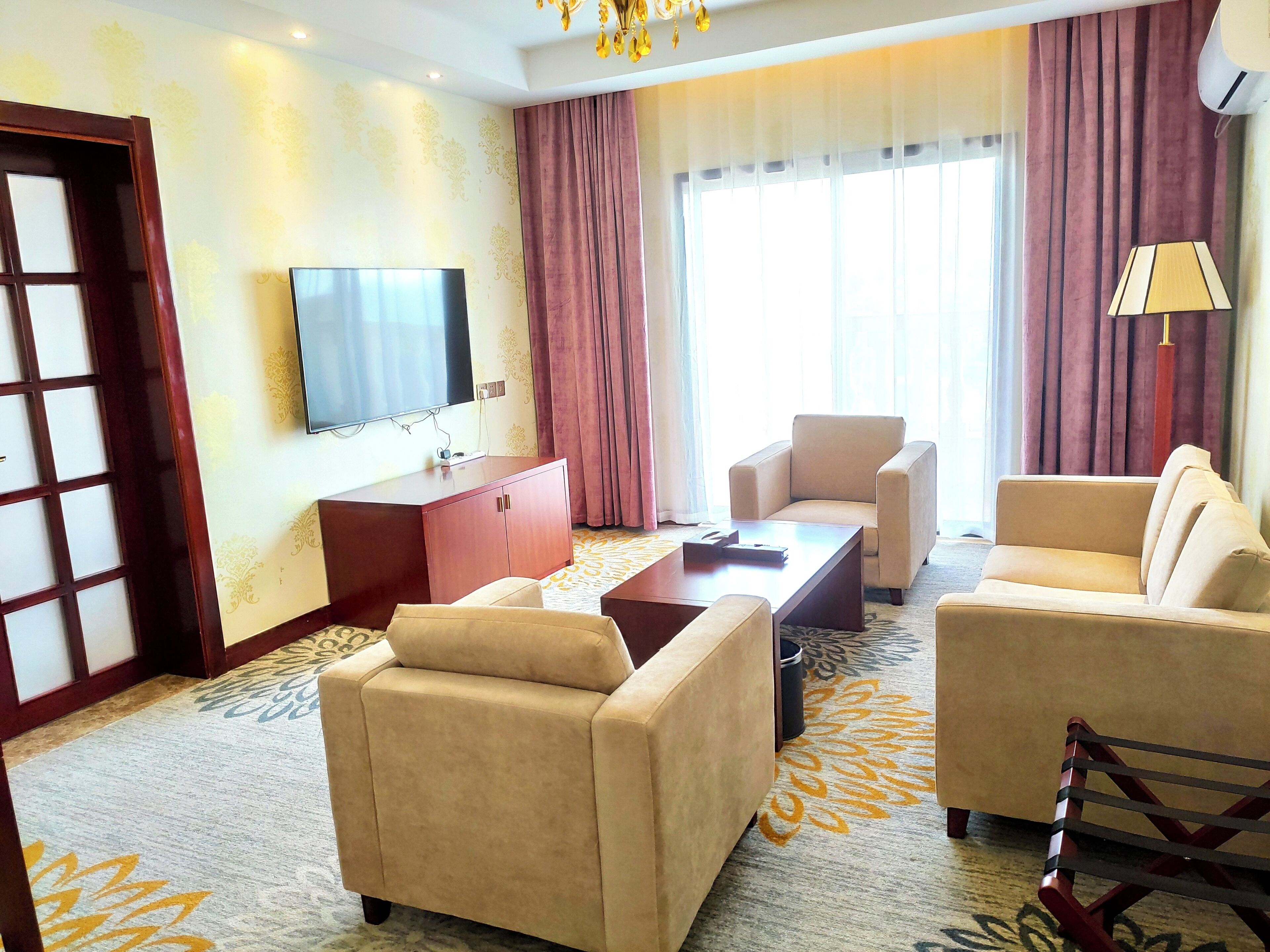 A hotel room with a flat-screen TV, a wooden cabinet, two beige sofas, a coffee table, and a patterned carpet.