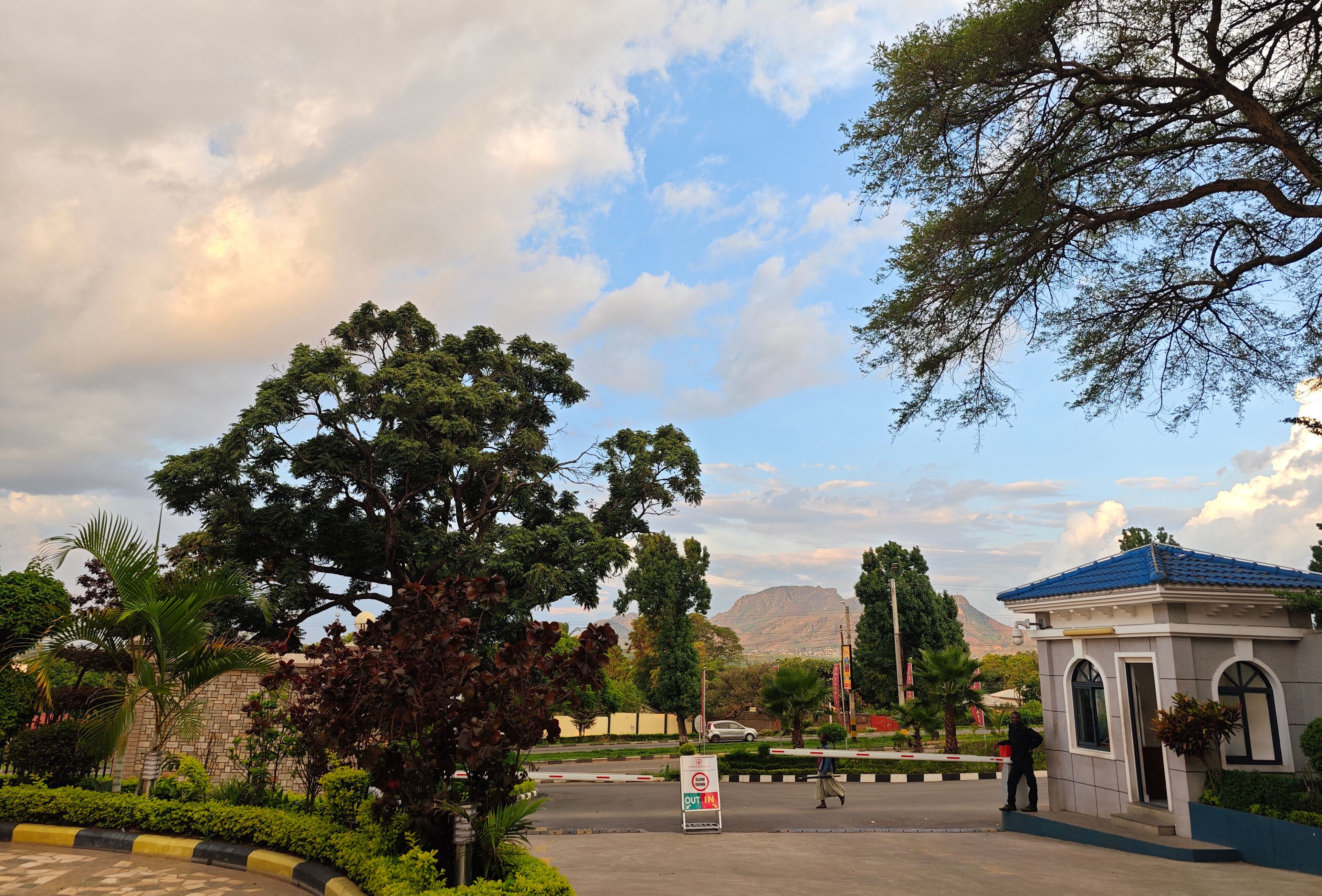 A gated entrance with a sign, a small building with a blue roof, and a mountainous background.