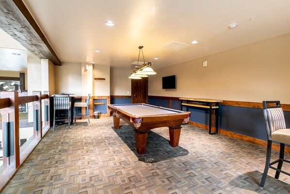 A pool table in a room with patterned carpet, wooden furniture, and a television on the wall.