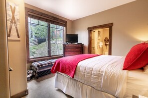 Une chambre à coucher avec un lit, un téléviseur posé sur une commode, une fenêtre donnant sur l’extérieur et une porte menant à une autre pièce.