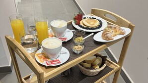 Un carrito de desayuno con diversos alimentos y bebidas, como panqueques, cruasanes y jugo, sobre un piso de baldosas.