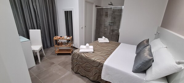 A hotel room with a bed, a bedside table, a small wooden shelf with toiletries, a shower area, and a view of the outside. - Anfitrite B&B (Tropea)