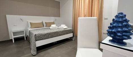 Une chambre d’hôtel moderne avec un lit soigneusement fait, une table de chevet blanche, une chaise blanche et un vase bleu décoratif.