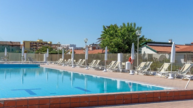 A swimming pool with lounge chairs and umbrellas, surrounded by a fence and buildings in the background. - Apartment in Porto Recanati with 4 bedrooms sleeps 8 (Porto Recanati)