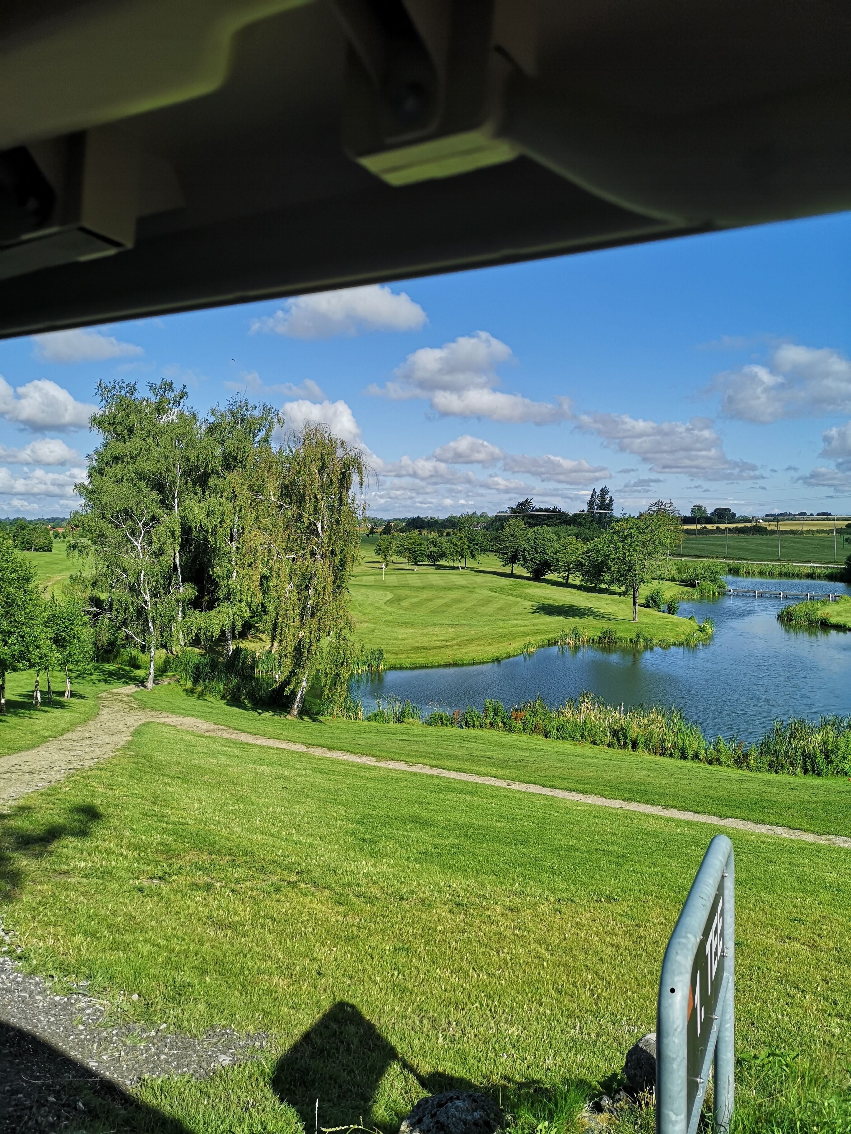 Vista desde el interior de un vehículo, con un parque cuidado que incluye un estanque, senderos y abundante vegetación.