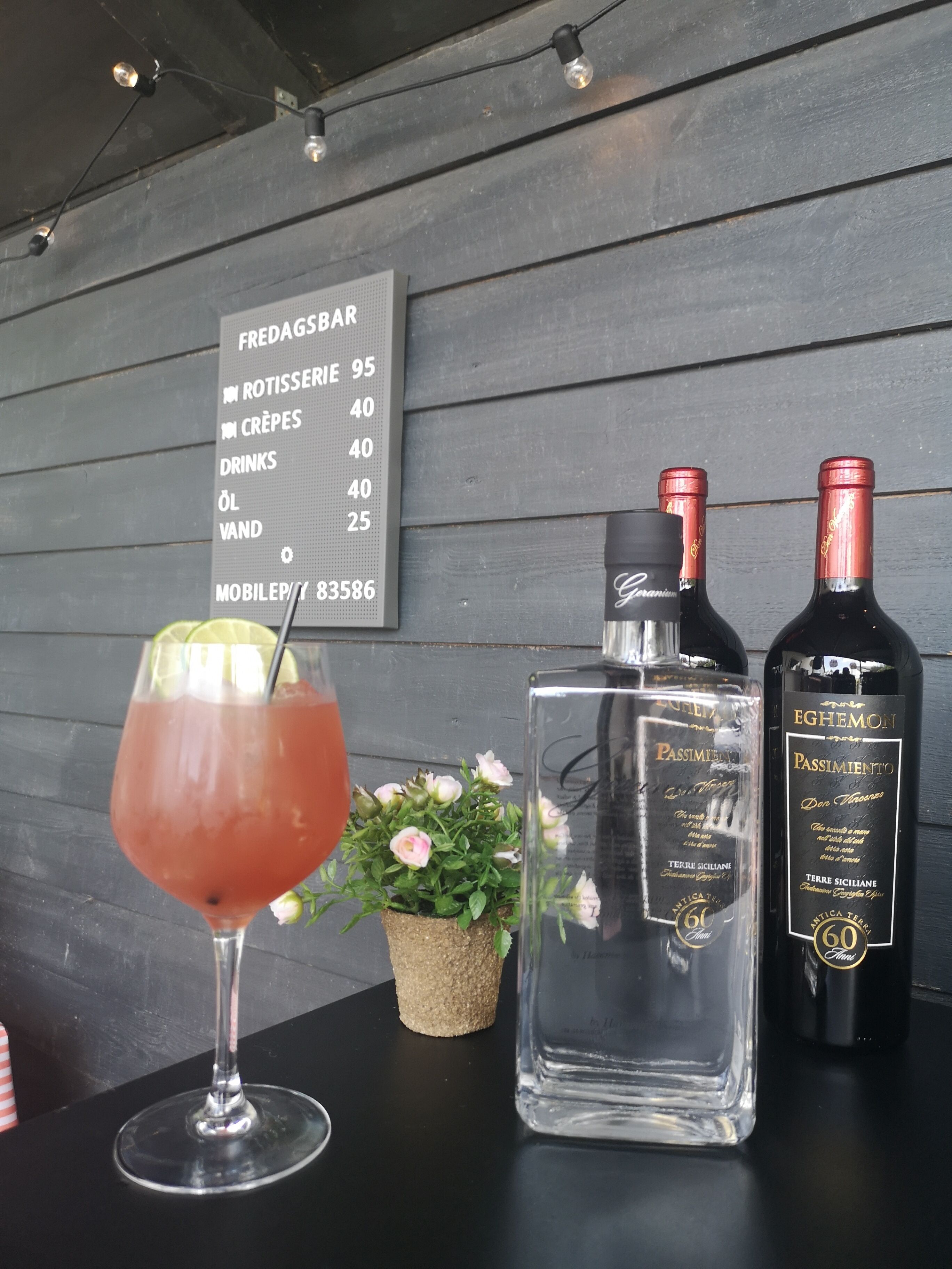 Een bar met een menubord waarop prijzen staan voor gerechten zoals kip van de draai, crêpes en drankjes. Op de tafel staan twee flessen Passimento wijn en een glas rode cocktail.