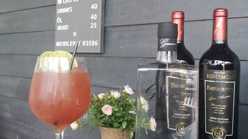 Een bar met een menubord waarop prijzen staan voor gerechten zoals kip van de draai, crêpes en drankjes. Op de tafel staan twee flessen Passimento wijn en een glas rode cocktail.