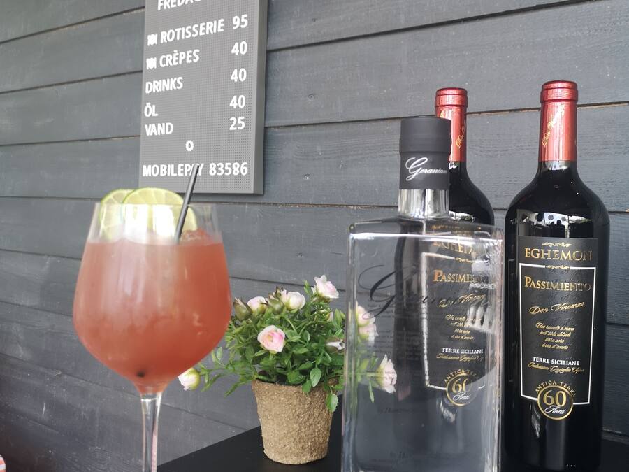 A bar area with a menu board listing prices for items like rotisserie, crepes, and drinks. There are two bottles of Passimento wine and a glass of red cocktail on the table.