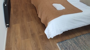 A bedroom with a wooden floor, a bed with white linens, a brown blanket, a hanging clothes rack, a framed picture on the wall, and a bedside lamp. - MidtGolf Guest house (Regstrup)