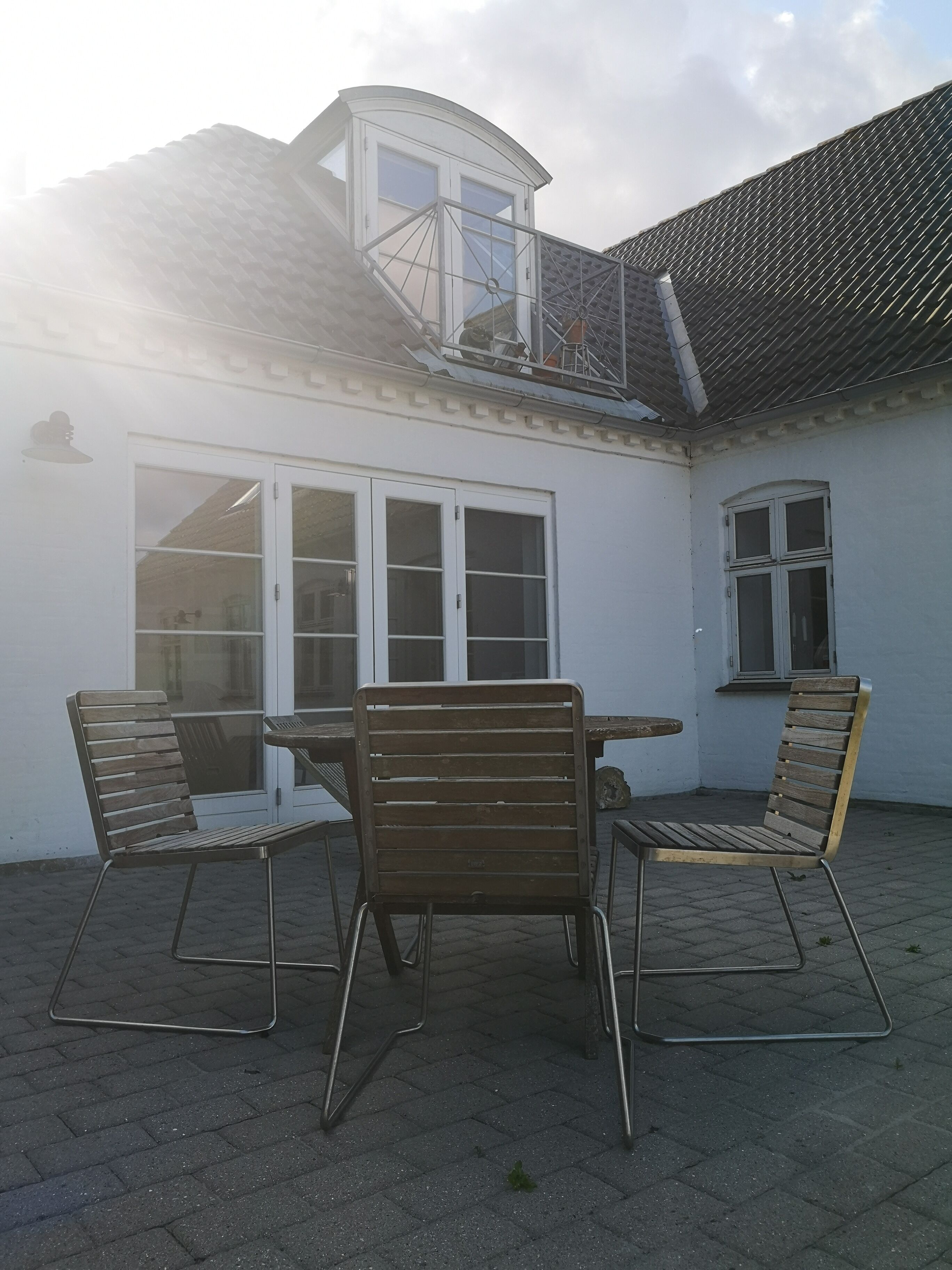 Pihalla on patio, jossa on pöytä ja neljä tuolia, pieni parveke sekä valkoinen rakennus, jossa on suuret ikkunat ja ovi.