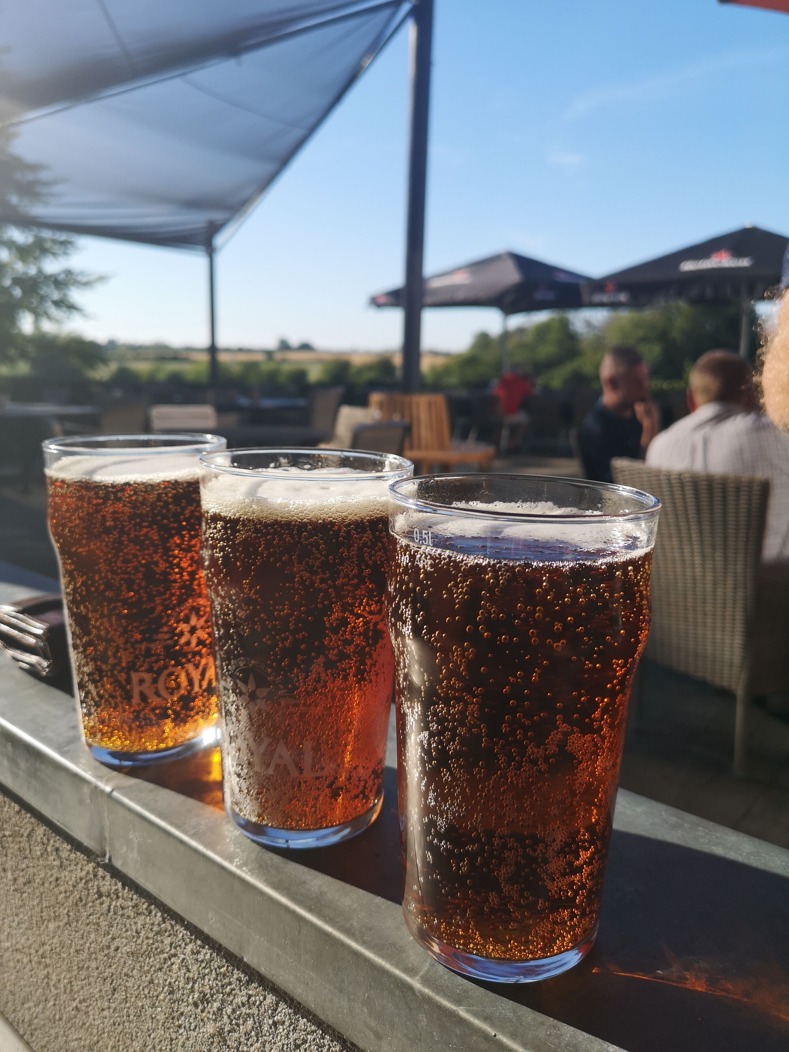 Three glasses of beer on a table with a clear view of the outdoors, including a covered area and people in the background.