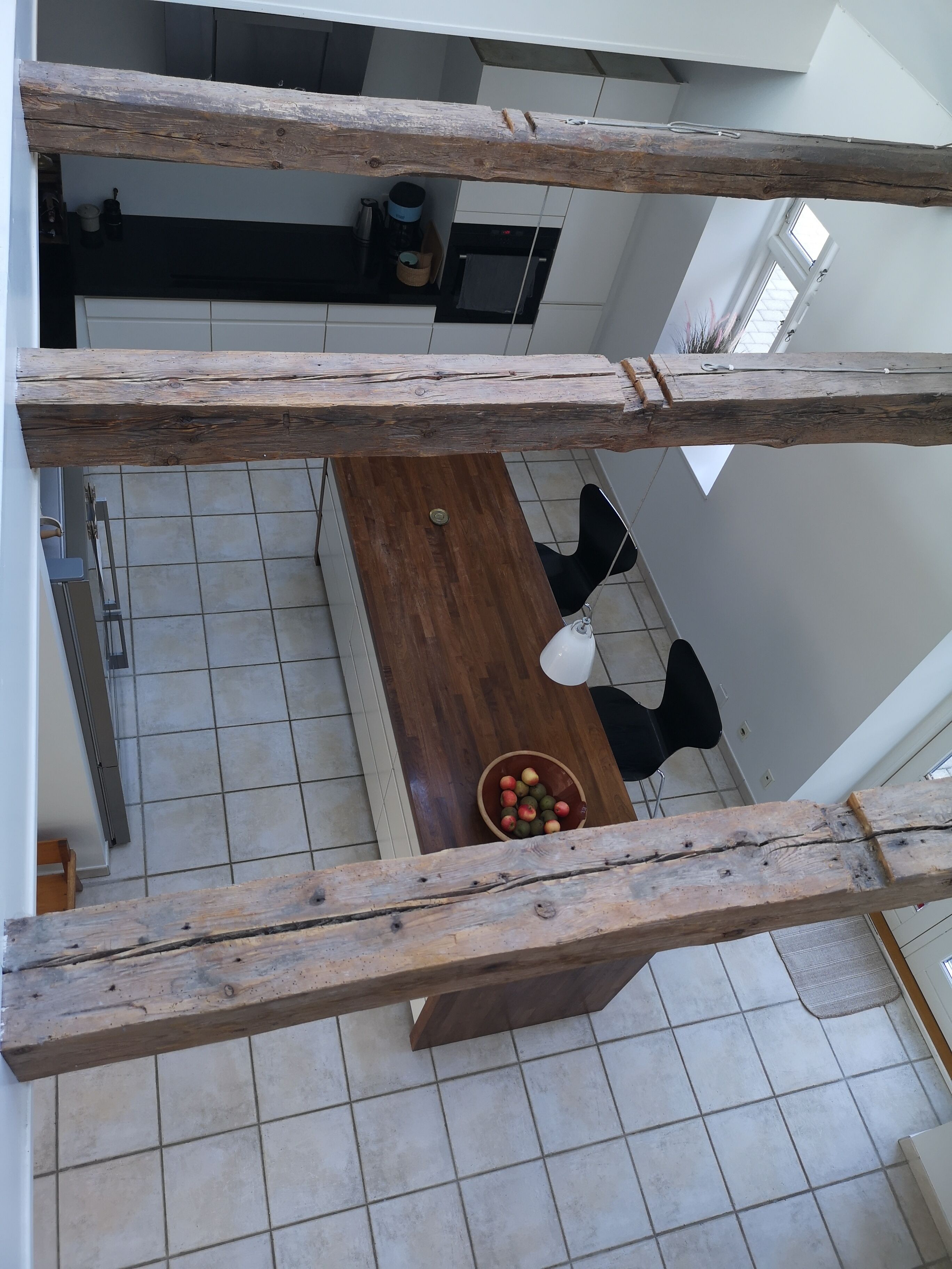 A high-ceilinged room with a wooden table and chairs, a bowl of fruit, and a view of a kitchen area.