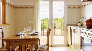 A dining area with a table set for four, a kitchen with a stove and a pot, and a sliding glass door leading to a balcony with a view of trees. - CA SA Lloritana - 6 pax, private pool, wifi free! (Sencelles)