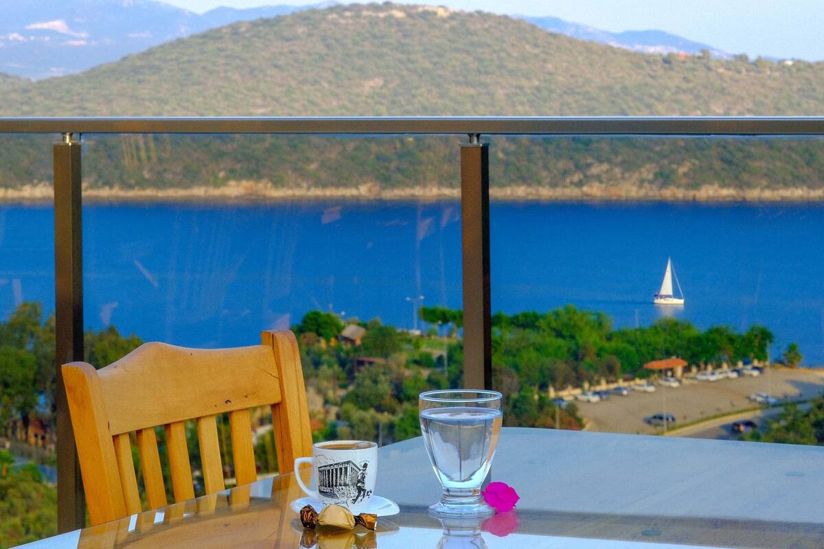A table with a glass of water, a mug, and chocolates, overlooking a coastal view with a sailboat and mountains.