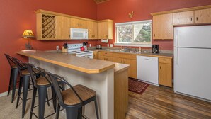 A kitchen with wooden cabinets, a white refrigerator, a microwave, and a coffee maker. There are four bar stools and a window with a view of trees.