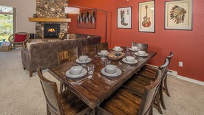 A dining area with a wooden table set for six, surrounded by wooden chairs. A stone fireplace with a mantel is visible in the background, and the room has red walls.