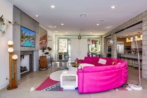 A modern living room with a vibrant pink sofa, a fireplace, and a kitchen in the background.