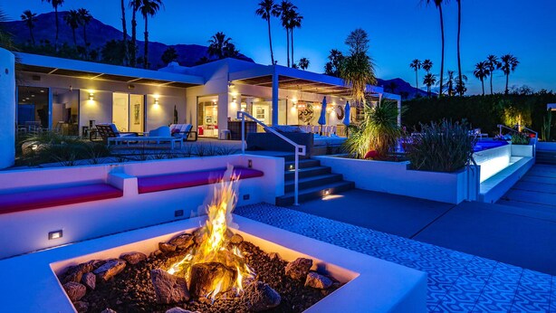 Een modern resort met een vuurplaats op de voorgrond, zitplaatsen buiten en palmbomen in de achtergrond.