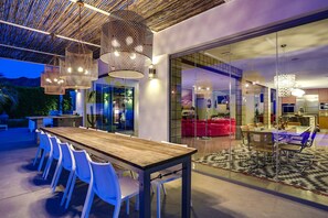 An outdoor dining area with a wooden table and white chairs, a glass wall separating it from the interior, and hanging woven light fixtures. - The Bird House - Swanky Poolside Paradise (Rancho Mirage)