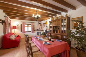 A dining area with a long table set for six, wooden beams on the ceiling, a red sofa, and a large potted plant.