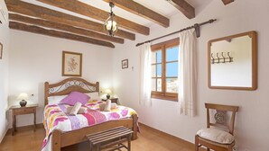 A bedroom with a wooden bed, a bench, a chair, a nightstand, a bedside table, a window with curtains, a mirror, and a wooden ceiling beam. - ASSUMPCIO. Pretty villa close to Pollensa (Pollensa)