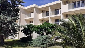 Um edifício com vários andares, varandas, aparelhos de ar condicionado e um gramado bem cuidado, com arbustos diversos e uma palmeira.