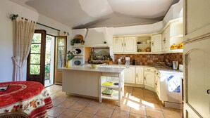 Una cocina con una isla central, una zona de comedor con una mesa roja, y vistas a un jardín a través de una puerta.