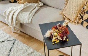 Ett vardagsrum med en soffa, ett litet svart bord med en blomsteruppsättning och en vit pläd.