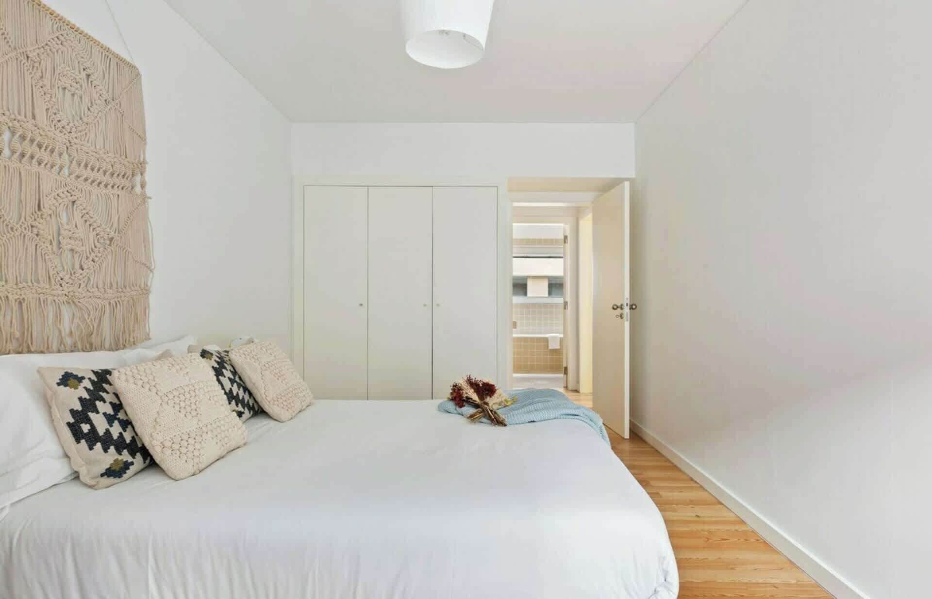 Una camera da letto con un letto, cuscini, un arazzo in macramè e una porta che conduce al bagno.