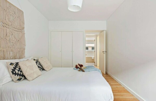 Une chambre à coucher avec un lit, des oreillers, une décoration murale en macramé et une porte donnant sur une salle de bain.