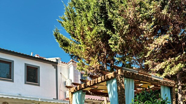 An outdoor seating area with wicker chairs and a wooden table with a cushion. A pergola with blue curtains is visible in the background. - My Dream Bozcaada (Bozcaada)