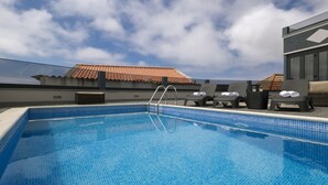 A clear blue pool with a tiled edge, two lounge chairs with towels, and a view of a building with a tiled roof. - ANGEL'S SUNSET T2 ESTRITO DA CALHETA (Estreito da Calheta)