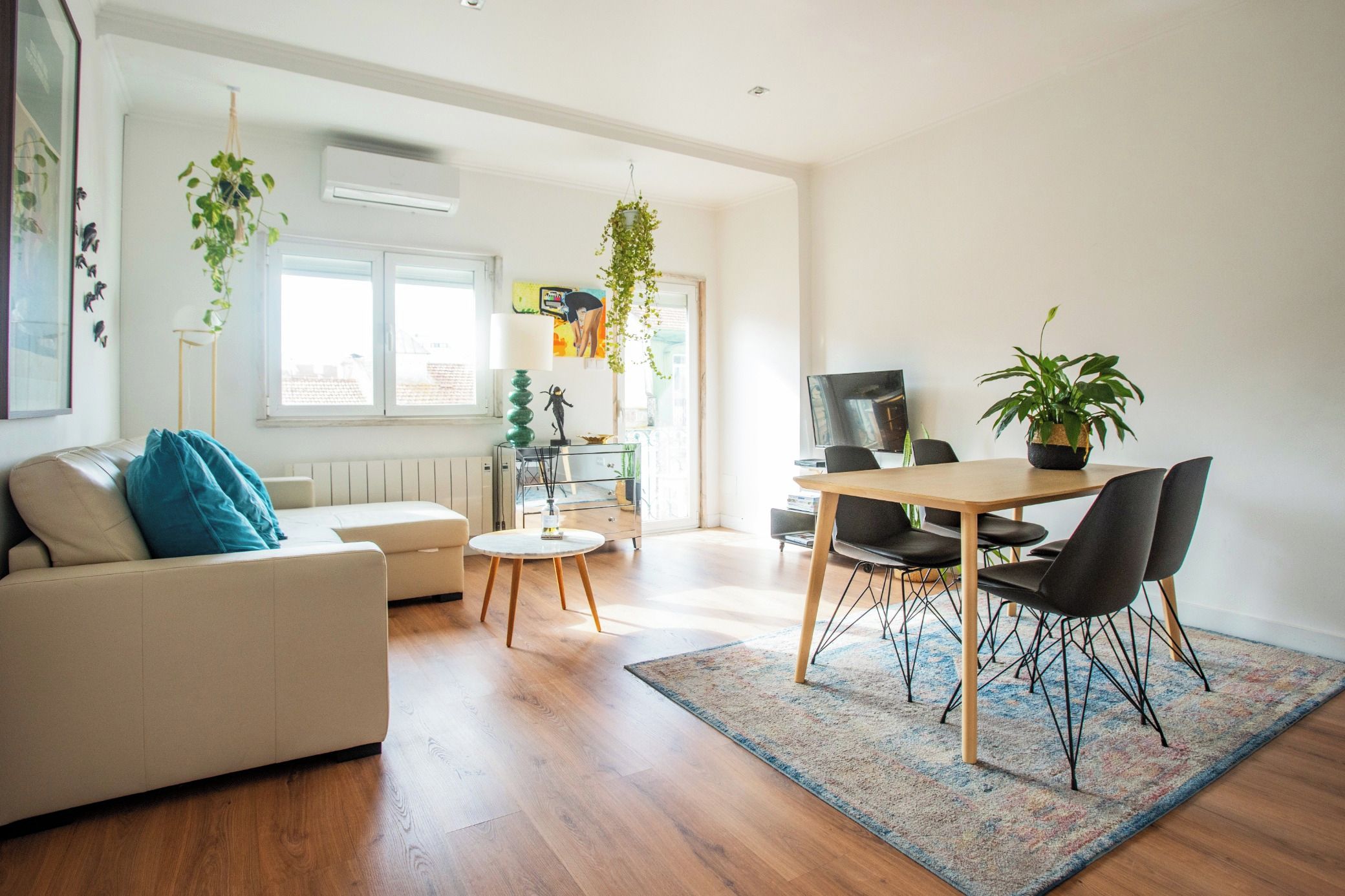 A modern living room with a sofa, a coffee table, a dining table with chairs, and a TV. There are plants and artwork on the walls.