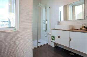 Ein modernes Badezimmer mit einer in Glas eingeschlossenen Dusche, einem weißen Waschtisch mit einem grünen Notausgangsschild und einem großen Spiegel über dem Waschbecken.