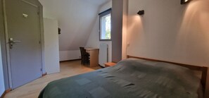 A small, well-lit bedroom with a single bed, a desk, and a chair. There is a window providing natural light and a door leading to another room.