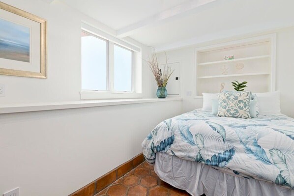 A bedroom with a large bed, a window, a shelf with decorative items, and a door. - Pink Venetian (lower unit) (Capitola)