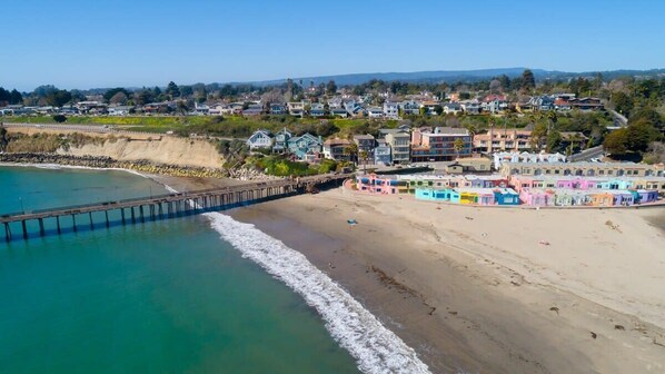 A coastal town with a pier extending into turquoise waters, colorful buildings along the beach, and a backdrop of residential houses.