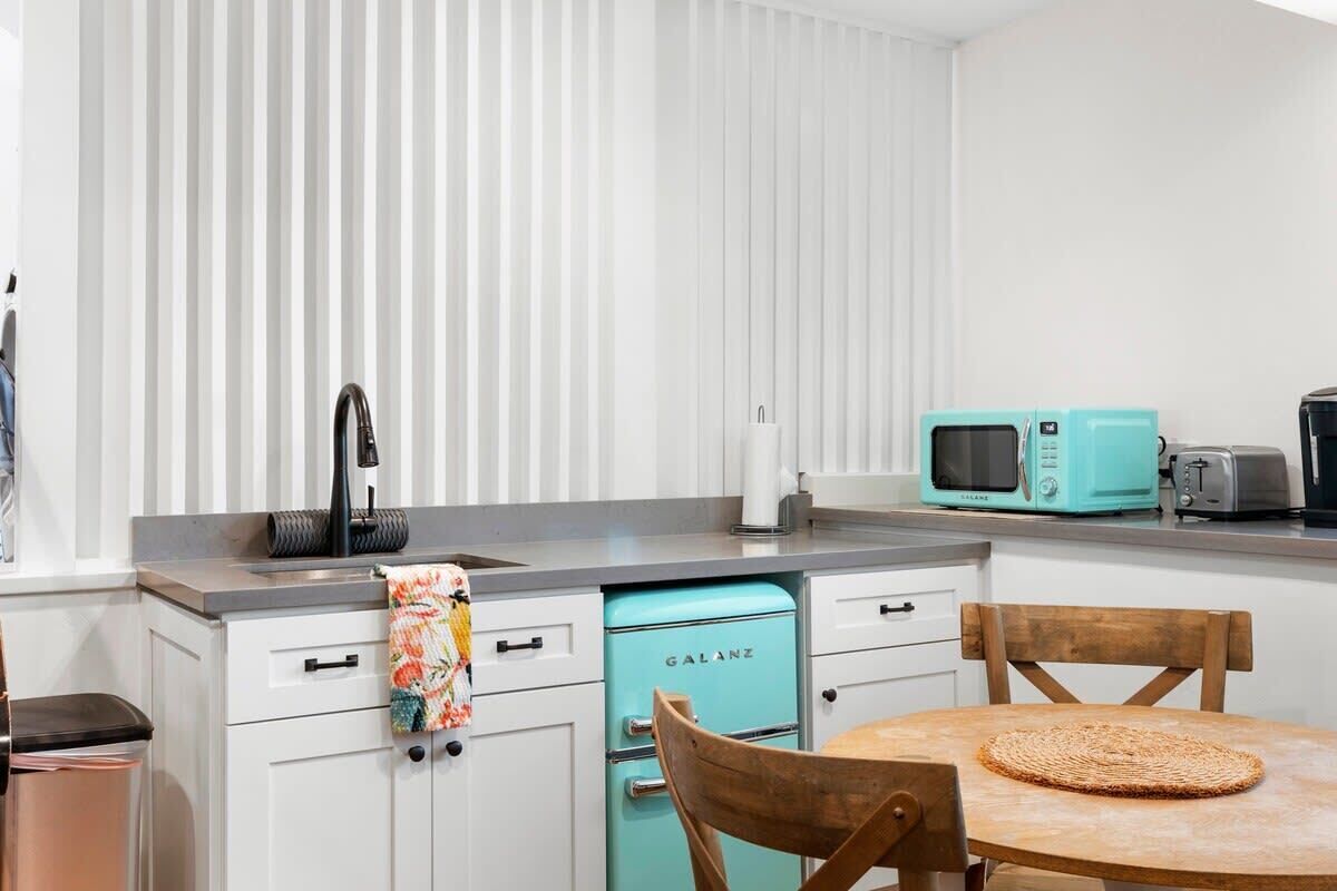 A kitchen with a sink, faucet, teal-colored dishwasher, microwave, toaster, and wooden chairs around a table.