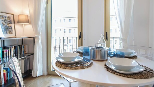 A dining area with a round table set for four, a bookshelf, and a view of the city through a large window.