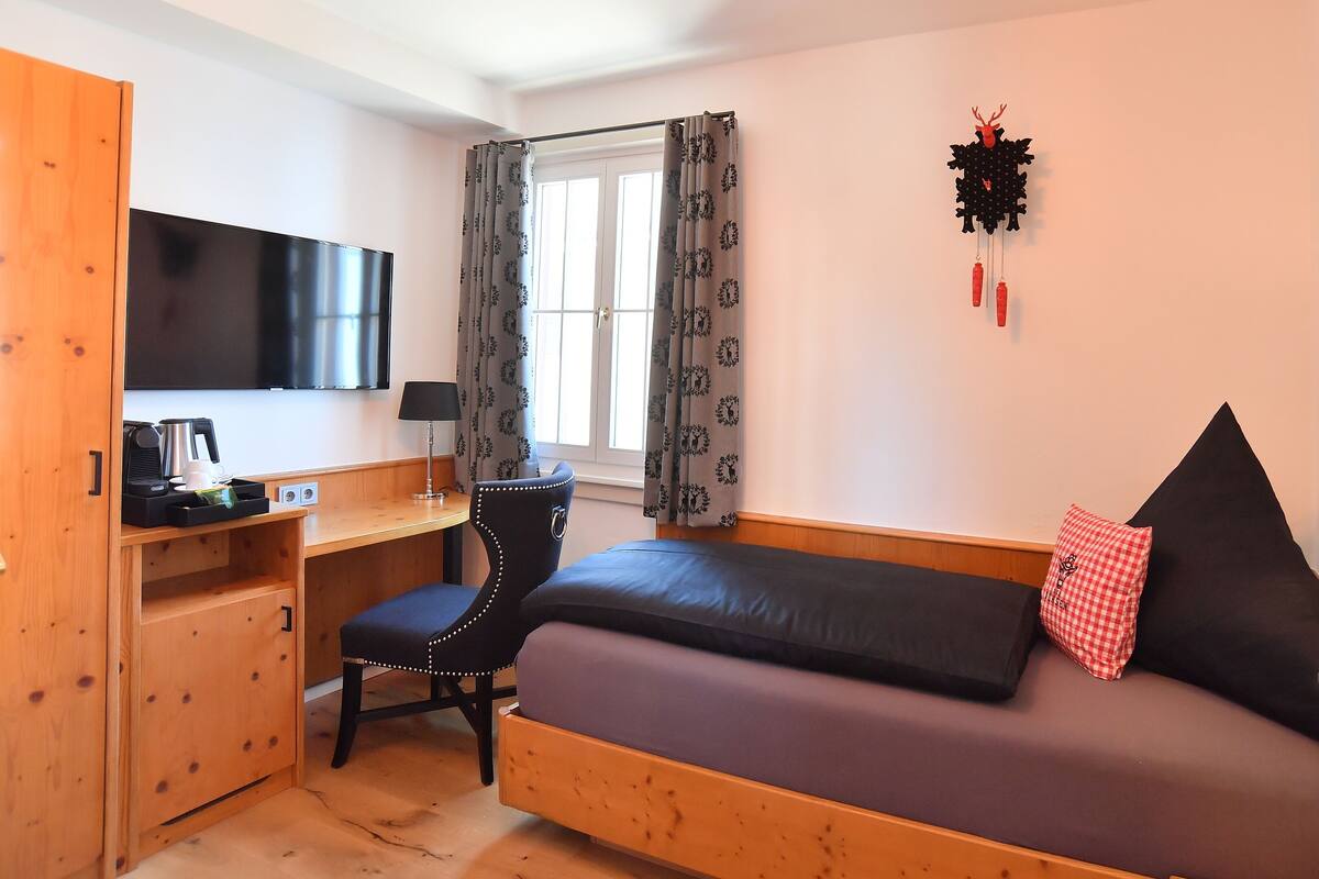 A bedroom with a bed, a desk, a chair, a TV, and a window with curtains.