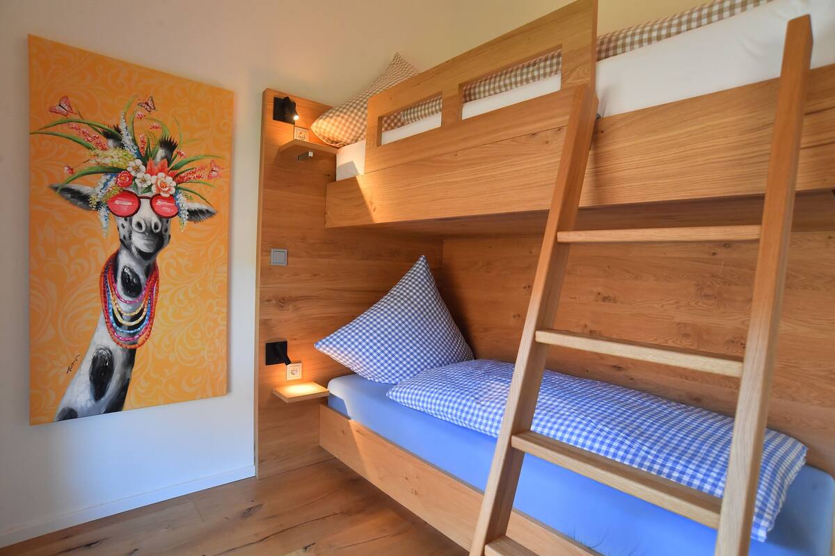 A bunk bed room with a checkered blue mattress, a wooden ladder, and a wall-mounted shelf.