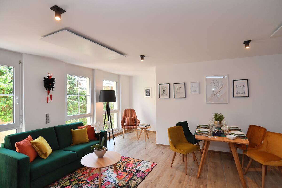 A modern living room with a green sofa, a dining table set for four, and a large window providing natural light.