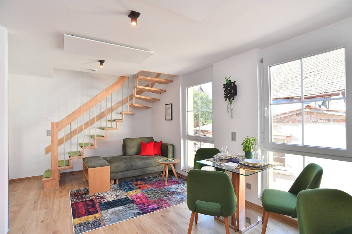 A modern living room with a staircase, a green sofa, a colorful rug, and a dining area with green chairs.
