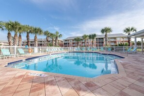 A swimming pool with a tiled deck, palm trees, and a building with balconies in the background.