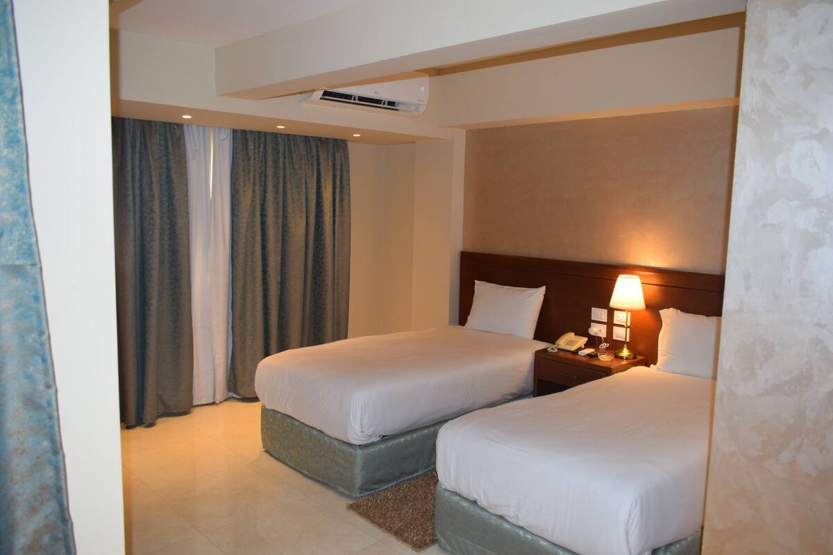 A hotel room with two beds, a nightstand with a lamp, and an air conditioning unit on the ceiling.