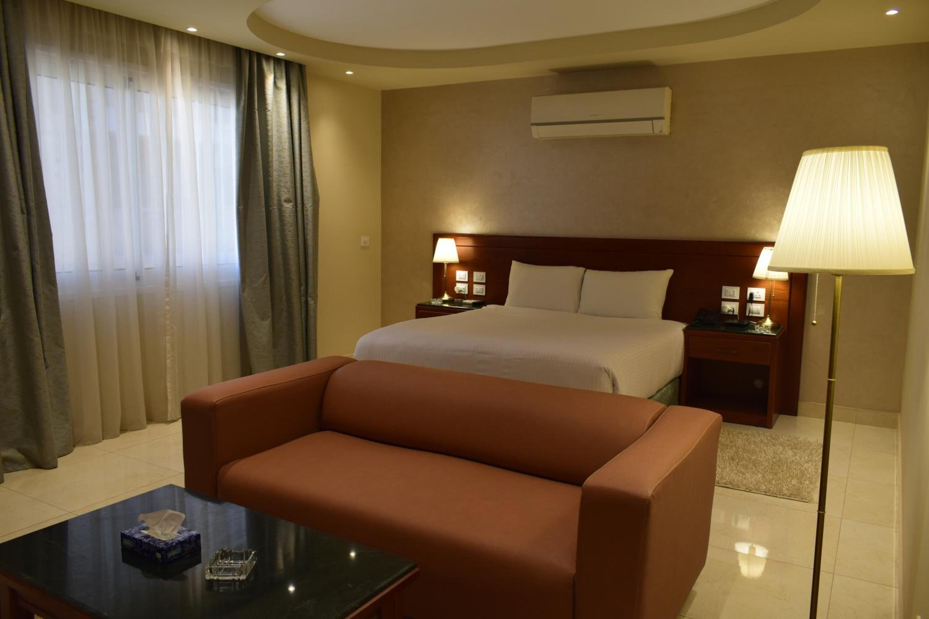 A hotel room with a bed, two bedside tables with lamps, a sofa, a coffee table, and a large window with curtains.